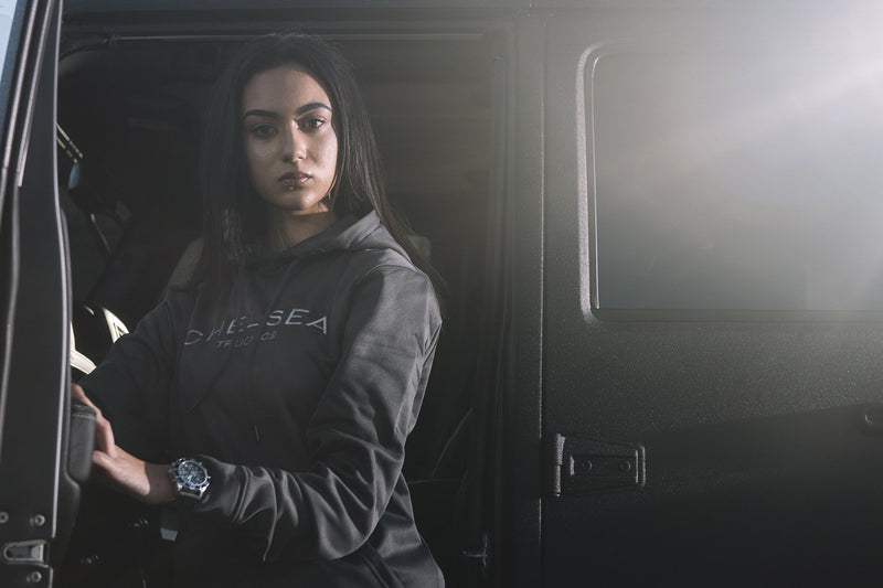 Female Model Wearing Chelsea Truck Co Hoodie - Steel Grey with Silver Embroidery Standing between Car Doors 